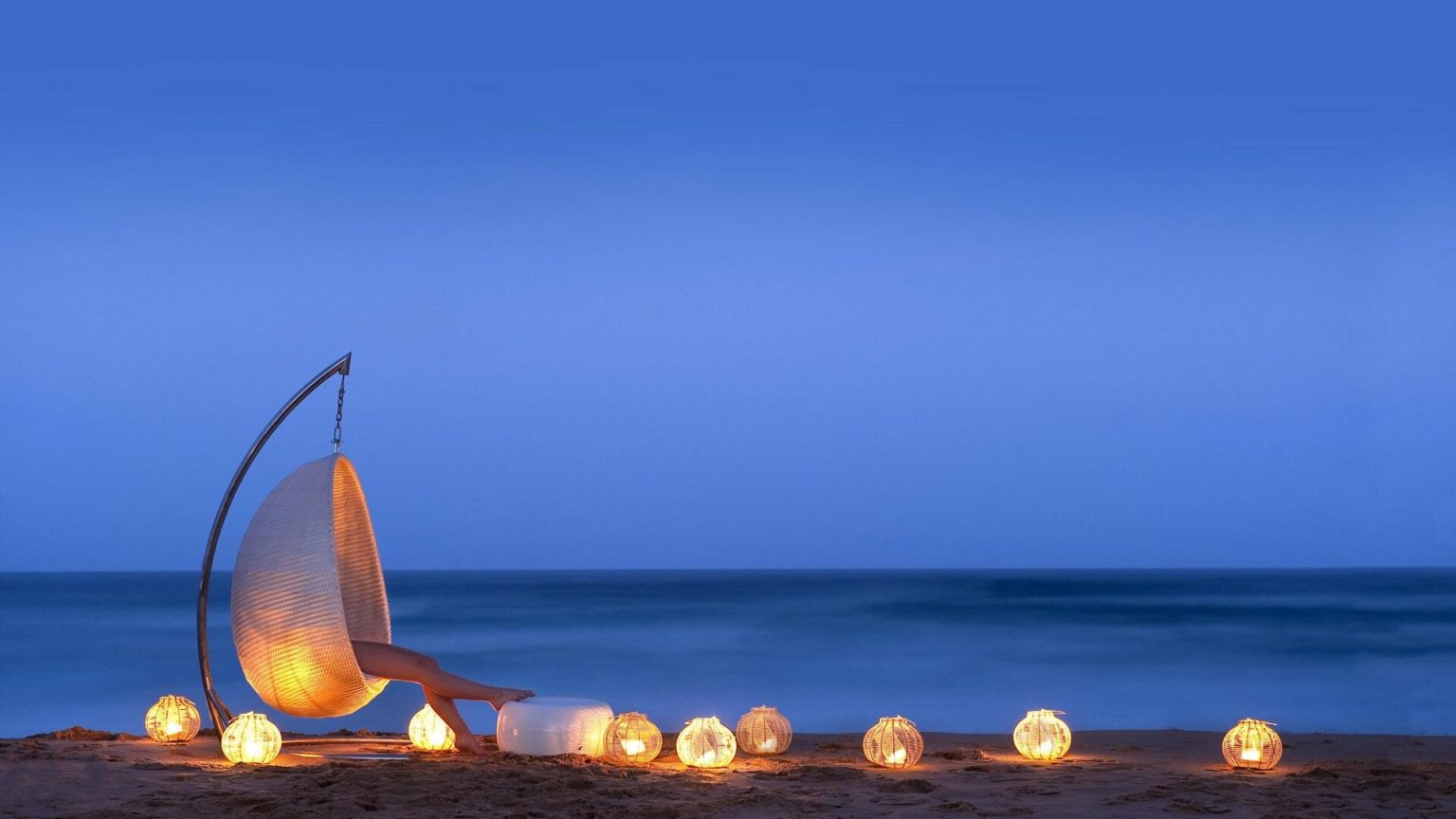 woman in a pearl shaped chair sits on the beach with lanterns at night