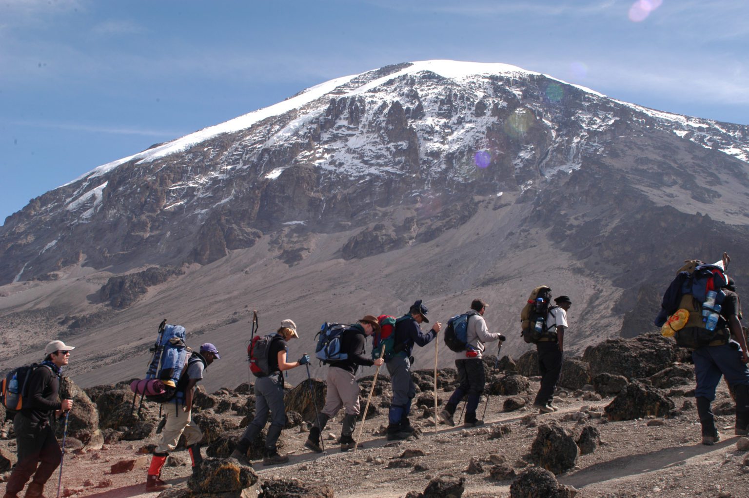 row of hikers with backpacks and walking sticks following the trail on Kilimanjaro with the snow-capped peak in the distance