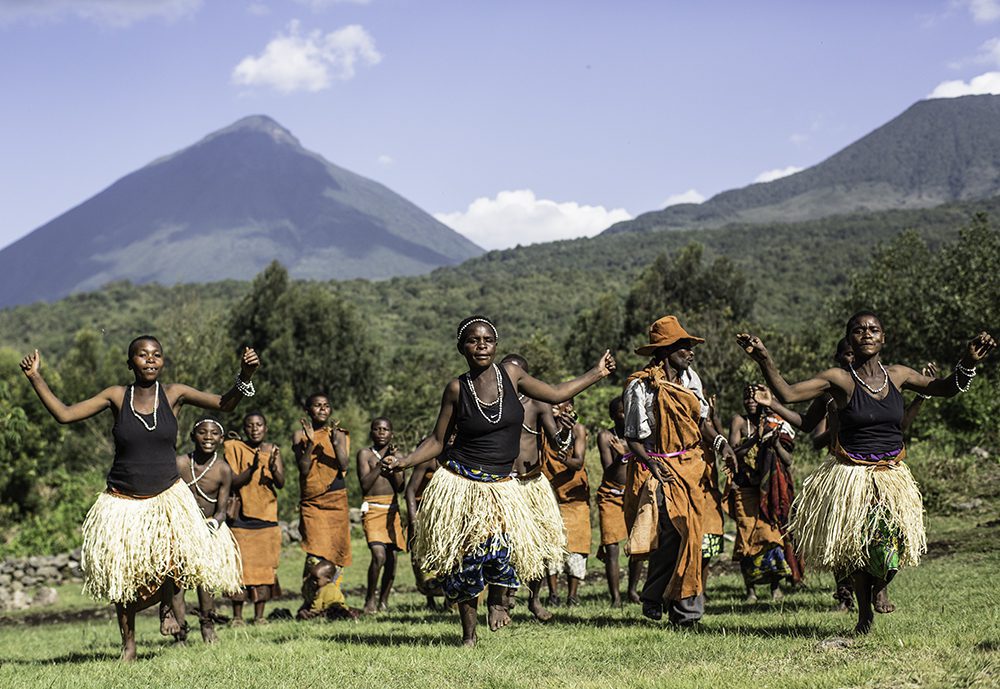 Batwa dancers in grass skirts and traditional clothing celebrate in front of the volcanoes