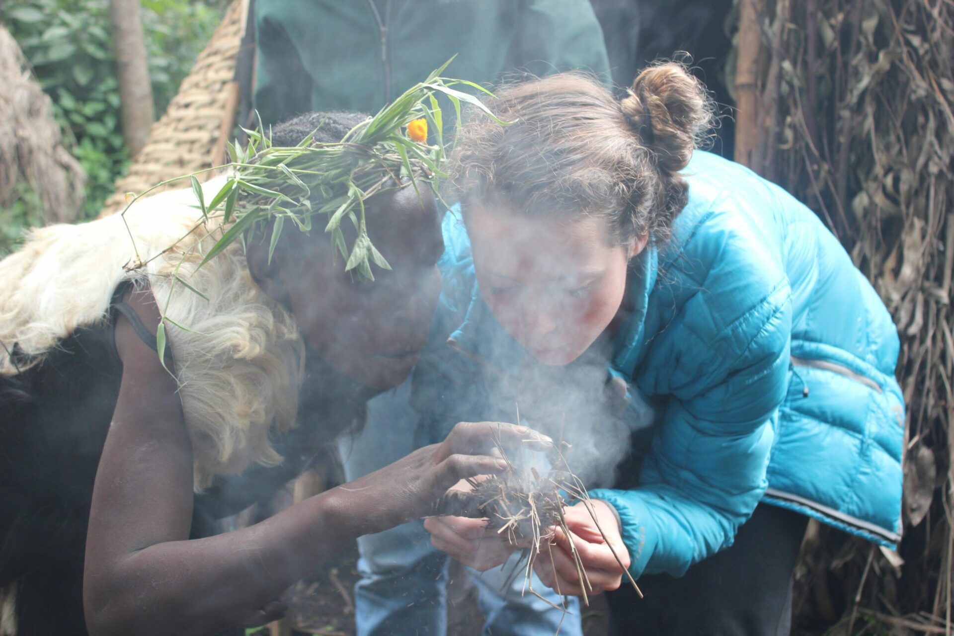 smoke is rising from sticks as a guest in a turquoise jacket helping a local Batwa make fire Uganda Safari