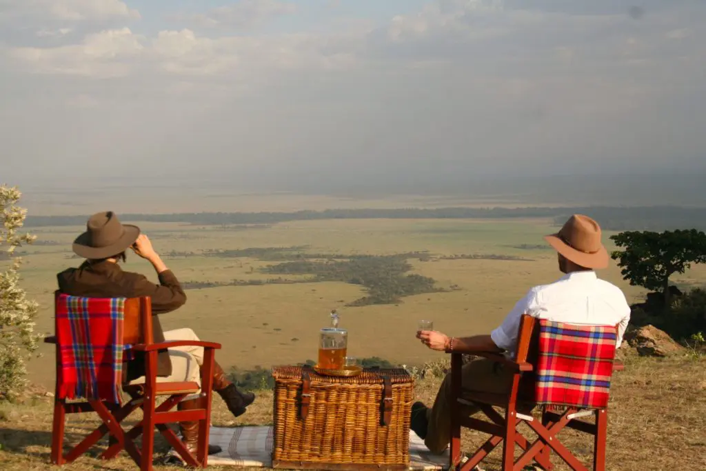 two people sit in chairs with blankets strewn over the back and enjoy a picnic overlooking the Maasai Mara