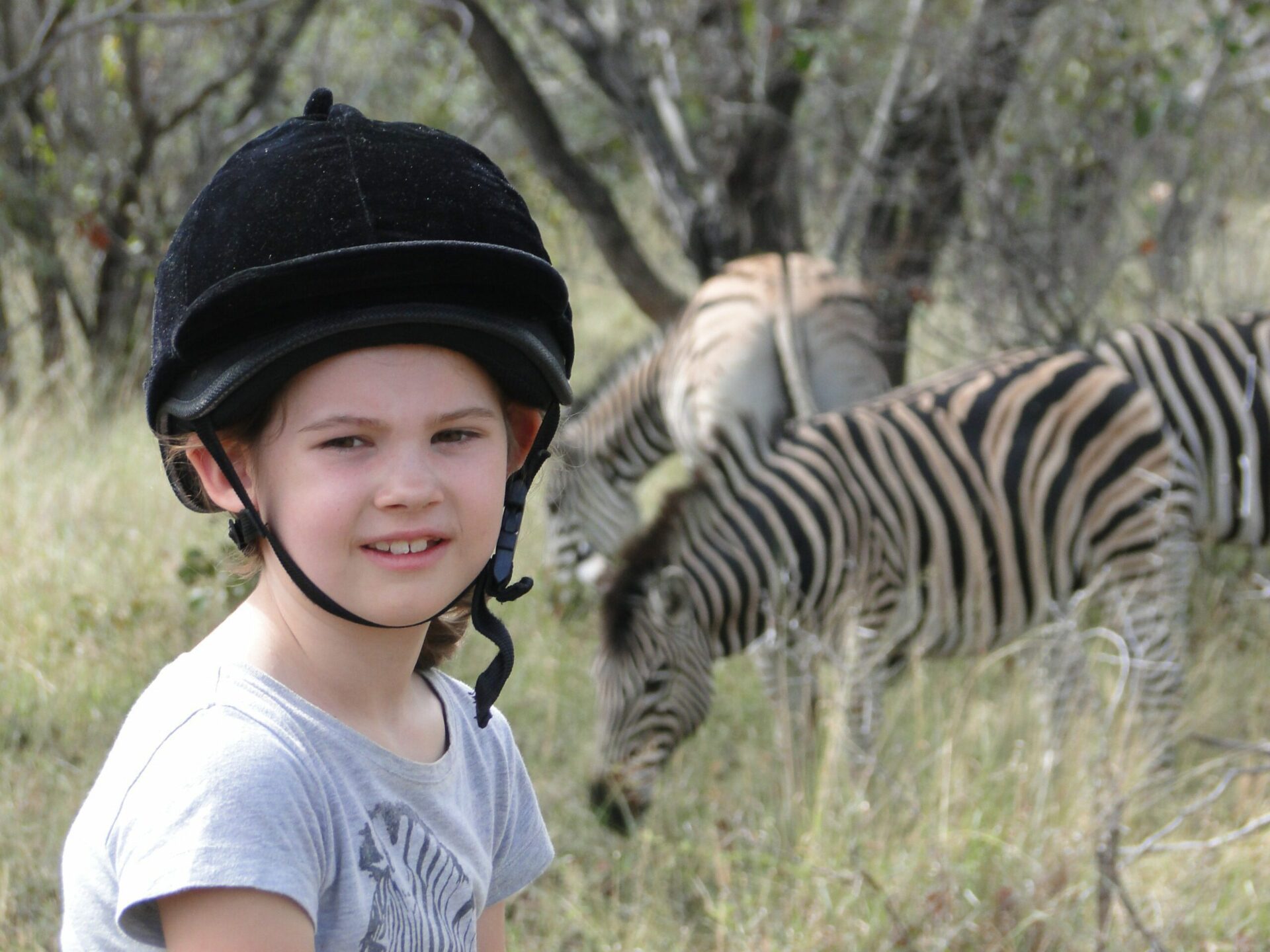 a young girl in a t-shirt wearing a riding helmet stops to pose for a photo in front of two zebra in the bush safari on horseback