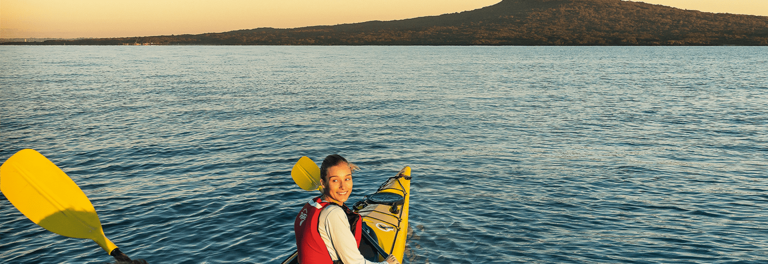 A happy young lady kayaks toward Rangitoto Island at dusk.
