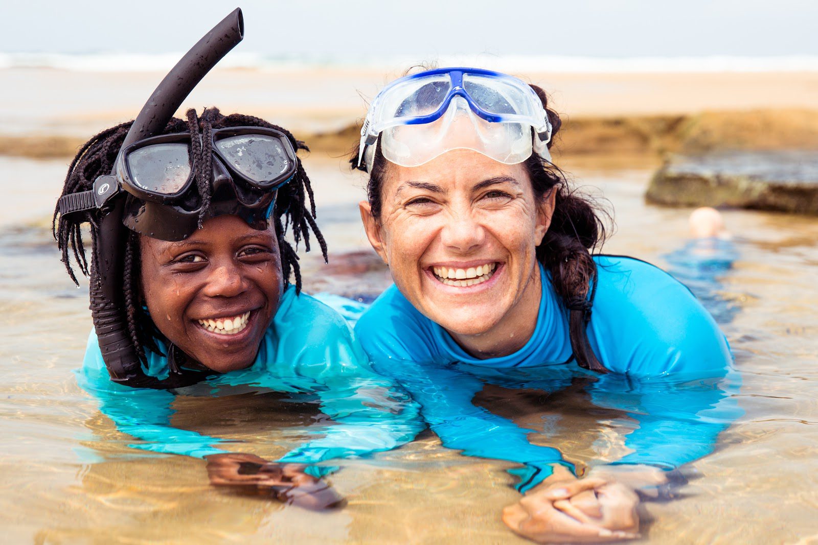 Hanli and a young girl lay in shallow, sandy waters, resting on their elbows & wearing blue rash guards, googles, & a snorkel