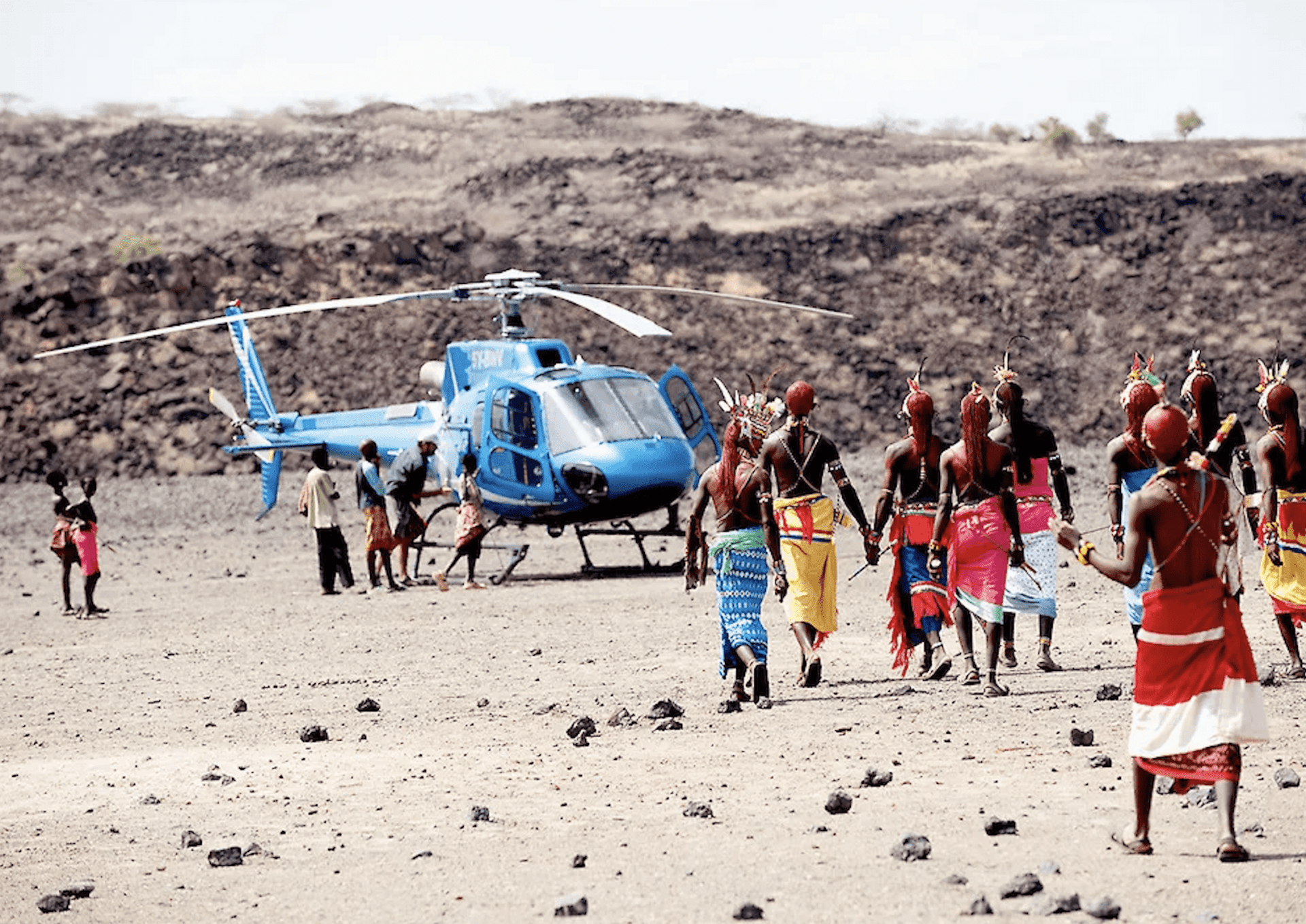 A crowd of people in colorful wraps and intricate headdresses gathers in a rocky landscape around a bright blue helicopter on this trip of a lifetime