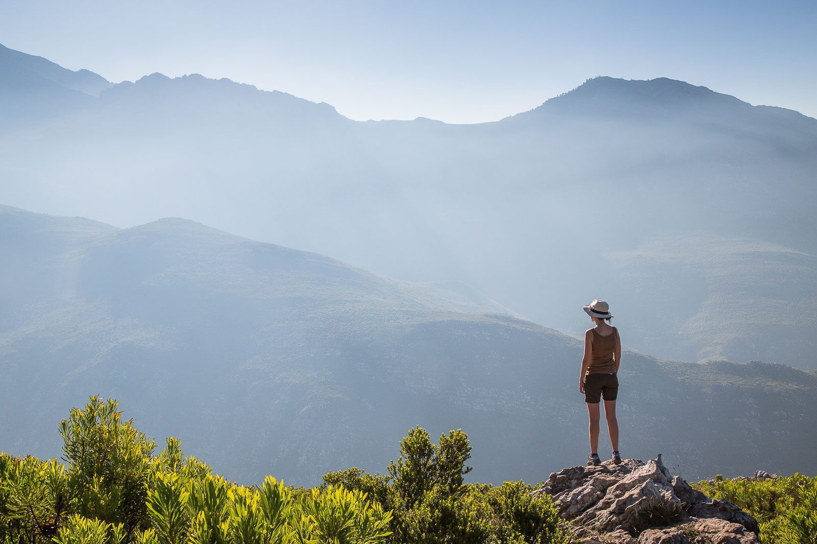 a woman hiker in a brimmed hat stands atop a rocky peak overlooking trees and towering, misty mountain ranges in the distance