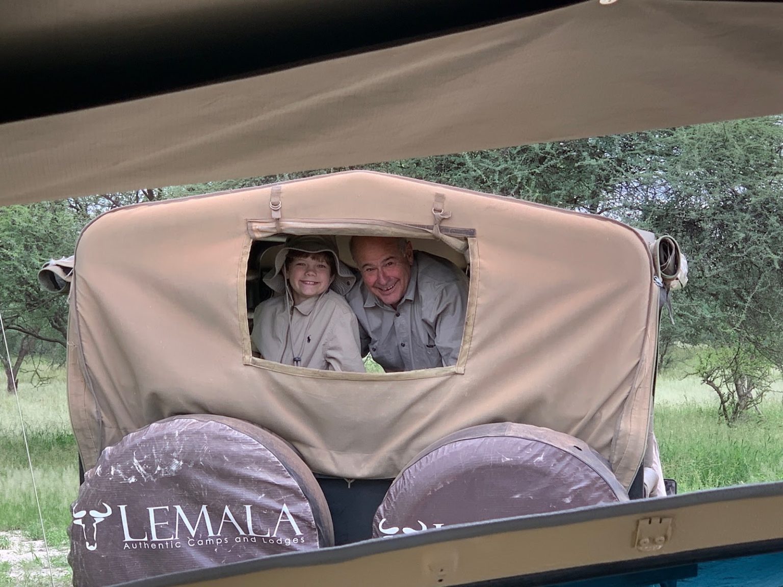 a young boy and his grandpa peer out the back window of a canvas-topped game drive vehicle, grinning