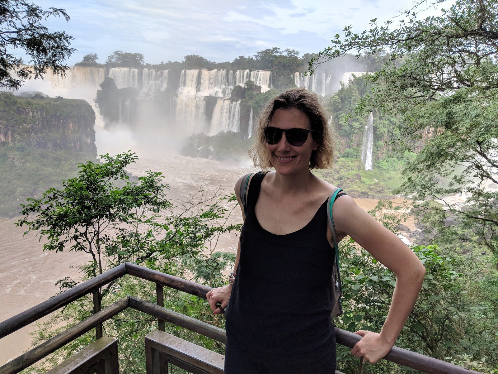 a woman in a black tank and black sunglasses smiles with a view of the misty Iguazu Falls behind her