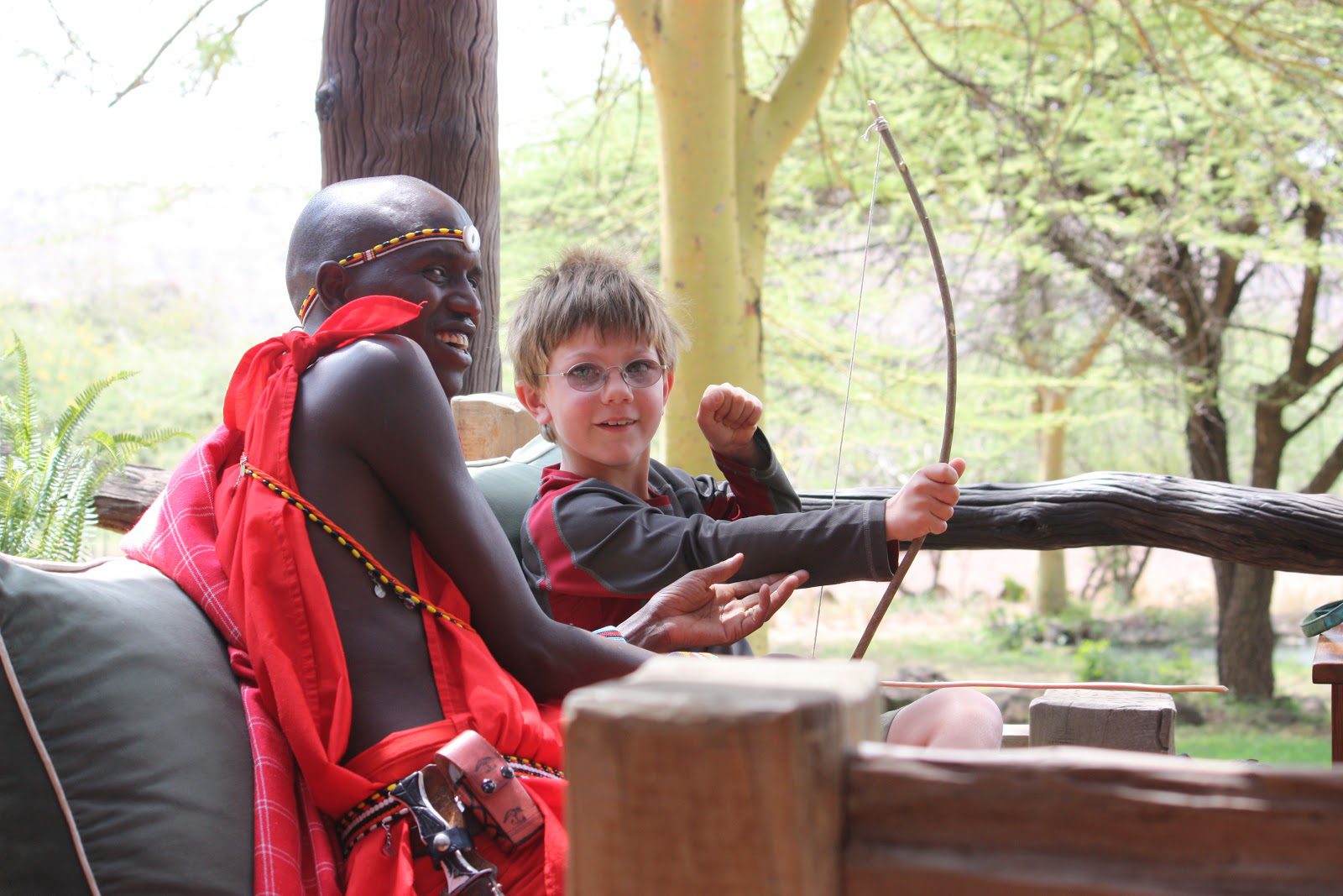 Kenya Luxury Safaris, A Young Boy Sits on a Sofa with a Bow and Arrow, Being Instructed by his Smiling Local Guide on its use