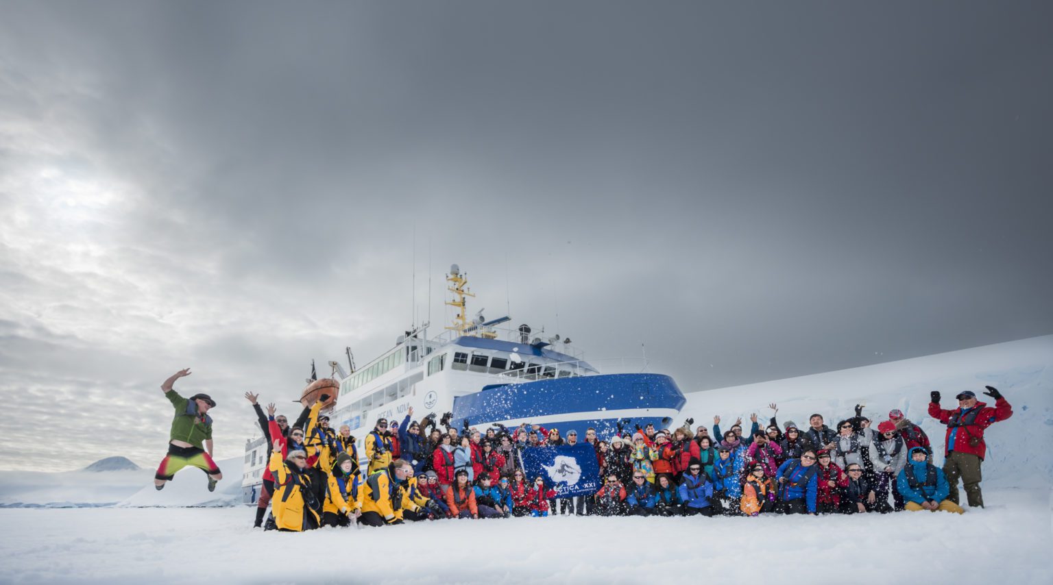 passengers and crew pose in front of their blue and white ice vessel celebrating their arrival to Antarctica