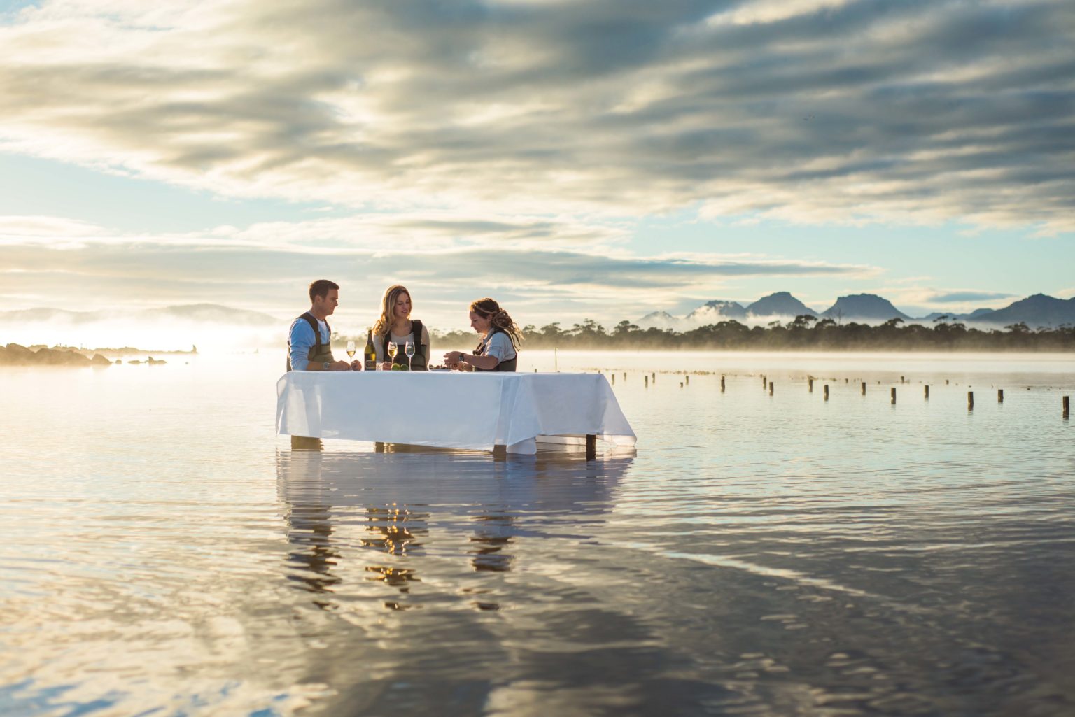 A couple and their guide sit in the shallow water at a table in an oyster farm where they're eating fresh oysters