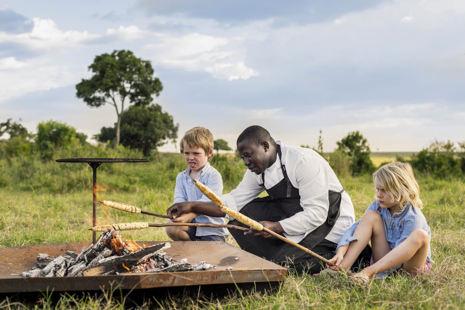 Chef at Angama Safari Camp helps two blonde children bake on skewers over an open fire