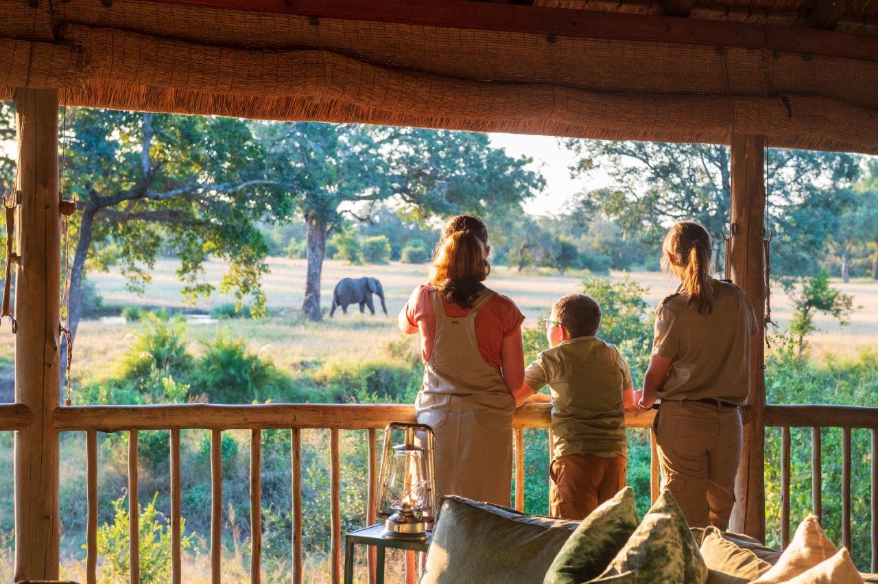 two women and a boy stand on a porch at the lodge to look at the elephant out in the plains