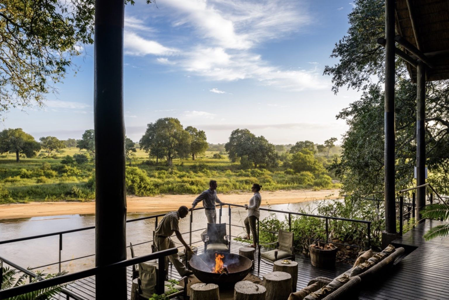 two people lean against a railing to talk while another adds wood to a fire pit on a balcony that overlooks the Sand River at Singita Ebony Lodge