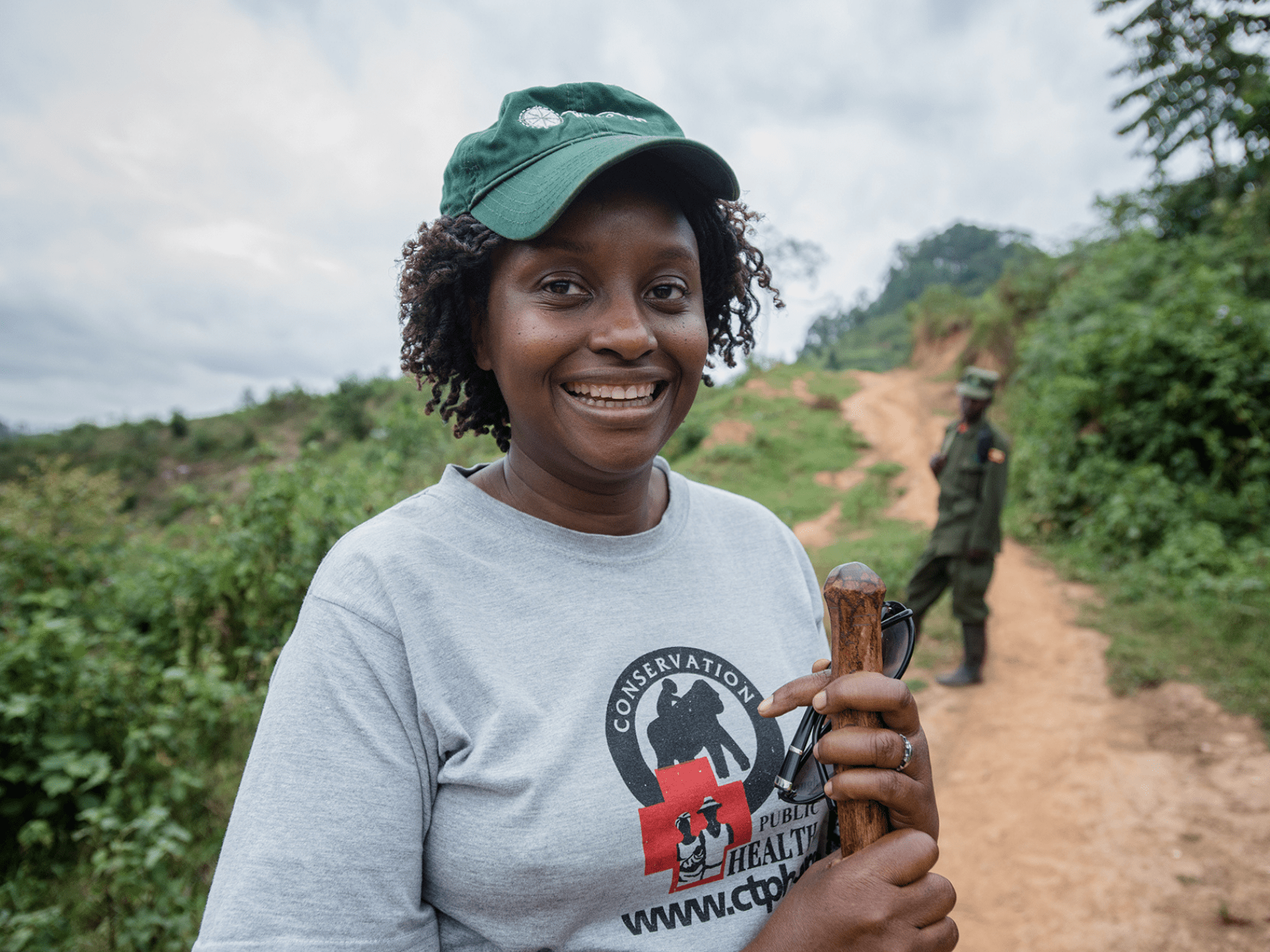 a smiling woman holding a wooden stick on a dirt road.