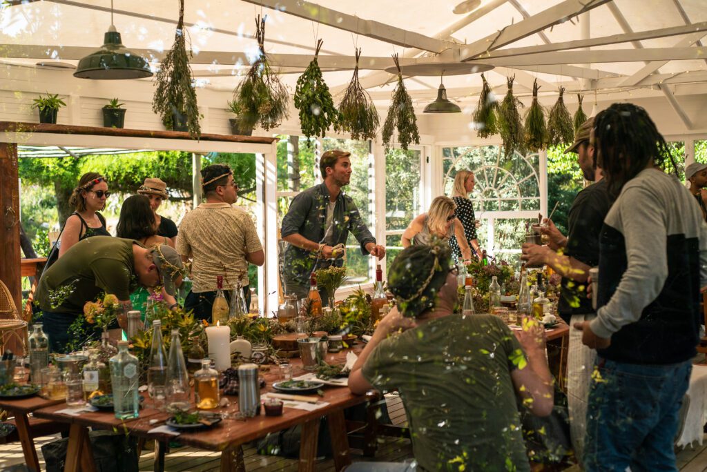 a Glasshouse filled with people communing around a beautifully dressed table with greenery hanging from the ceiling as well as decorating the table. 