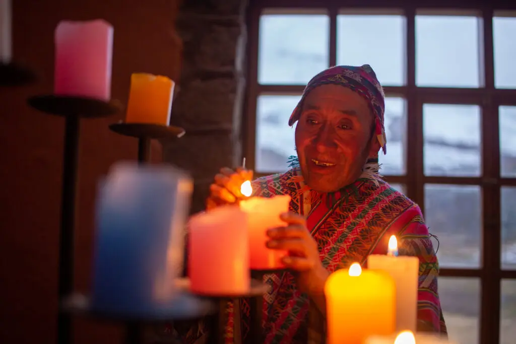 Quechua woman lighting candles with a window view of the Peruvian Andes blurred in the background. 
