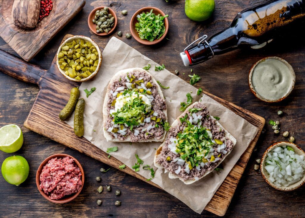Tight shot of an open face raw meat sandwich on a wooden board with limes, small bowls of chopped onion, sauce, and raw meat surrounding the sandwhich. 
