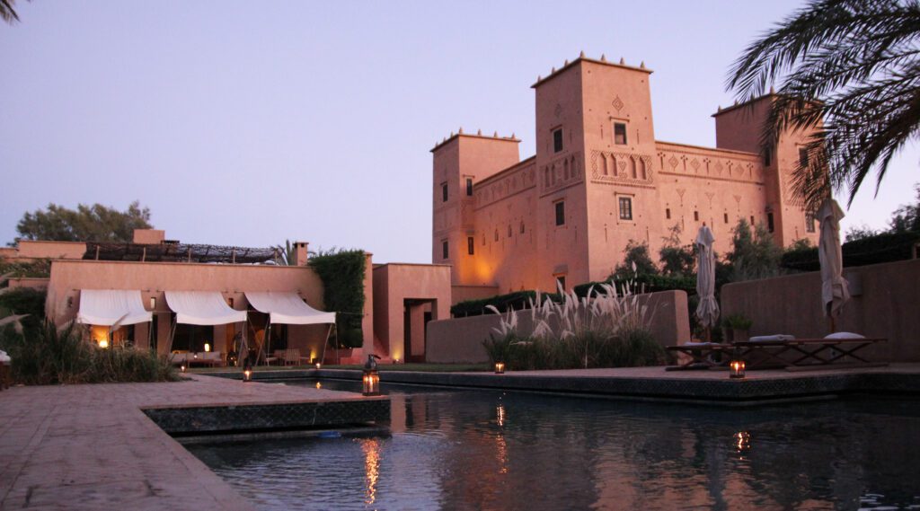 Dar Ahlam hotel grounds with the pool in the foreground and hotel behind it seated on the hem of the Moroccan desert and housed in a kasbah.