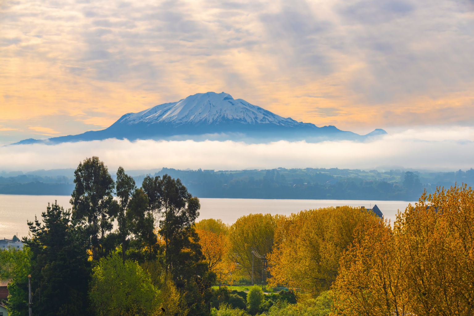 View of snow-capped Osorno volcano with a line of clouds halfway down with trees and the lake in the foreground