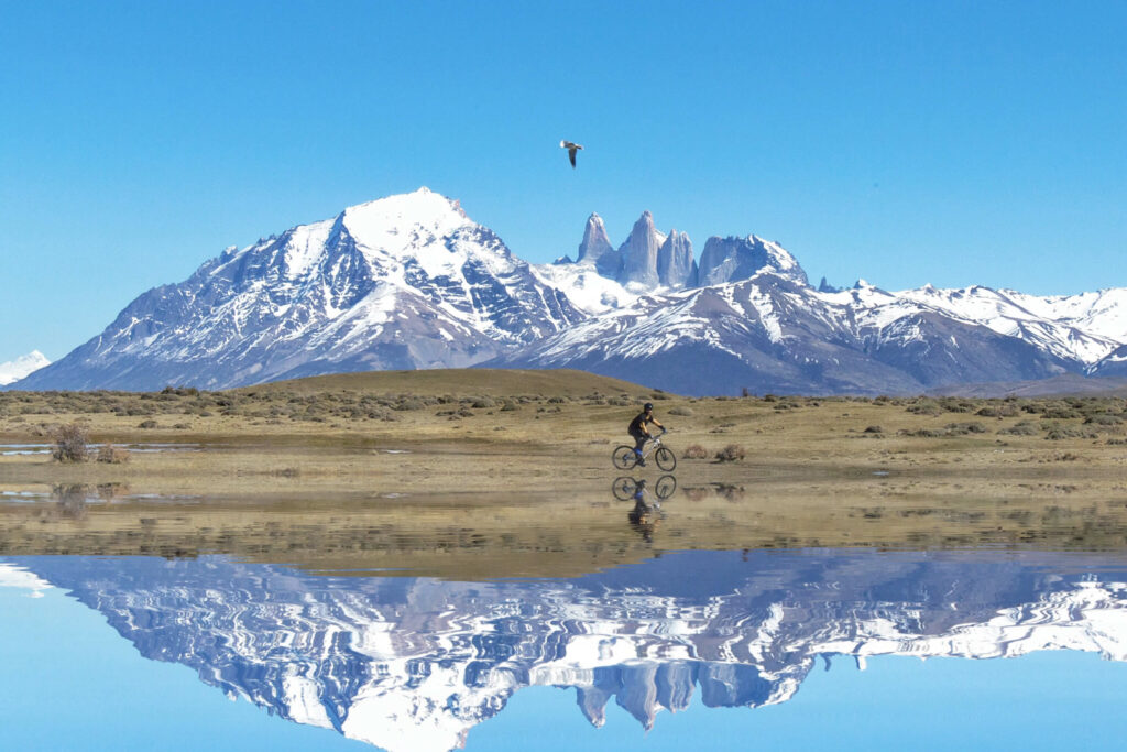 A cyclist rides near water with Torres del Paine in the background. 