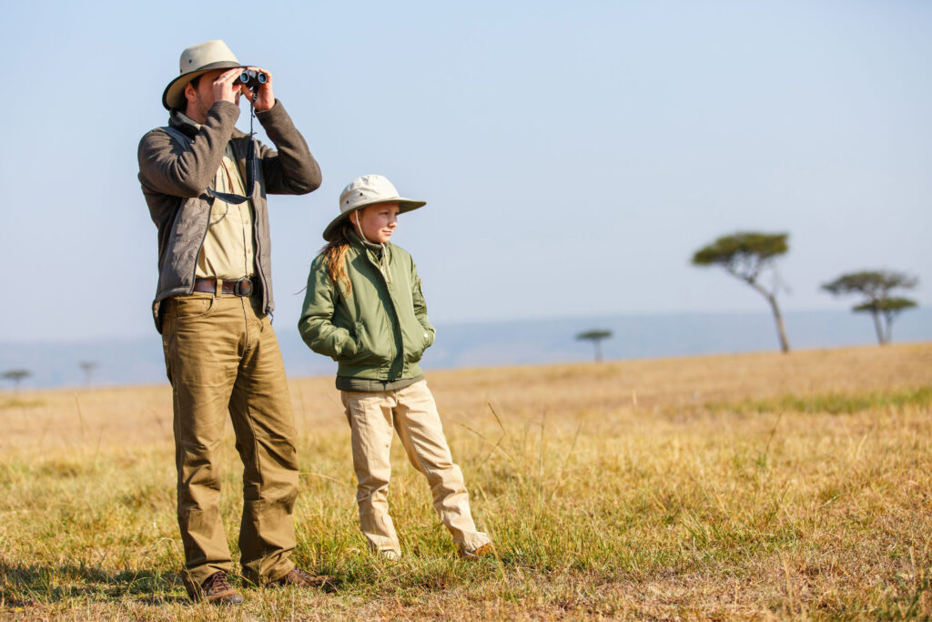 A man and young girl stand on the savannah dressed in safari clothing. 