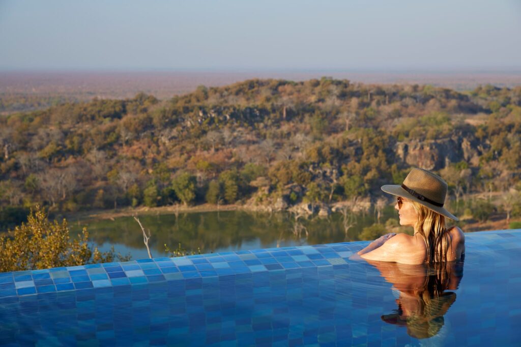 A woman in an infinity pool overlooking tree-covered hills and a small lake on safari.