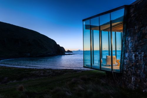 a corner room at Seascape features floor-to-ceiling windows with panoramic views of an ocean sunset and dramatic cliffs