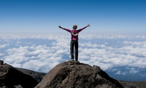 hiker with her arms outstretched and standing on a rock at the top of Mount Kilimanjaro towering above the clouds