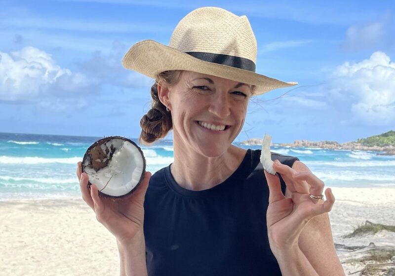 A woman holding a coconut and a straw hat on a beach.